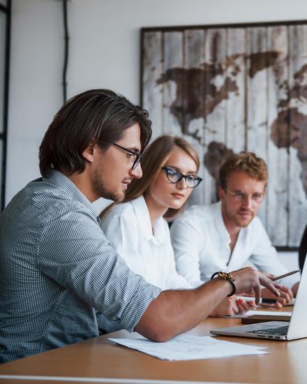 Drei Personen sitzen an einem Tisch und schauen auf einen Laptop, eine vierte Person steht im Hintergrund vor einer Wand mit einem großen Bild