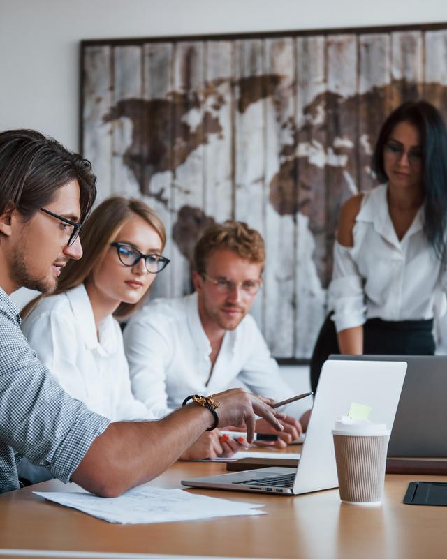 Drei Personen sitzen an einem Tisch und schauen auf einen Laptop, eine vierte Person steht im Hintergrund vor einer Wand mit einem großen Bild Drei Personen sitzen an einem Tisch und schauen auf einen Laptop, eine vierte Person steht im Hintergrund vor einer Wand mit einem großen Bild