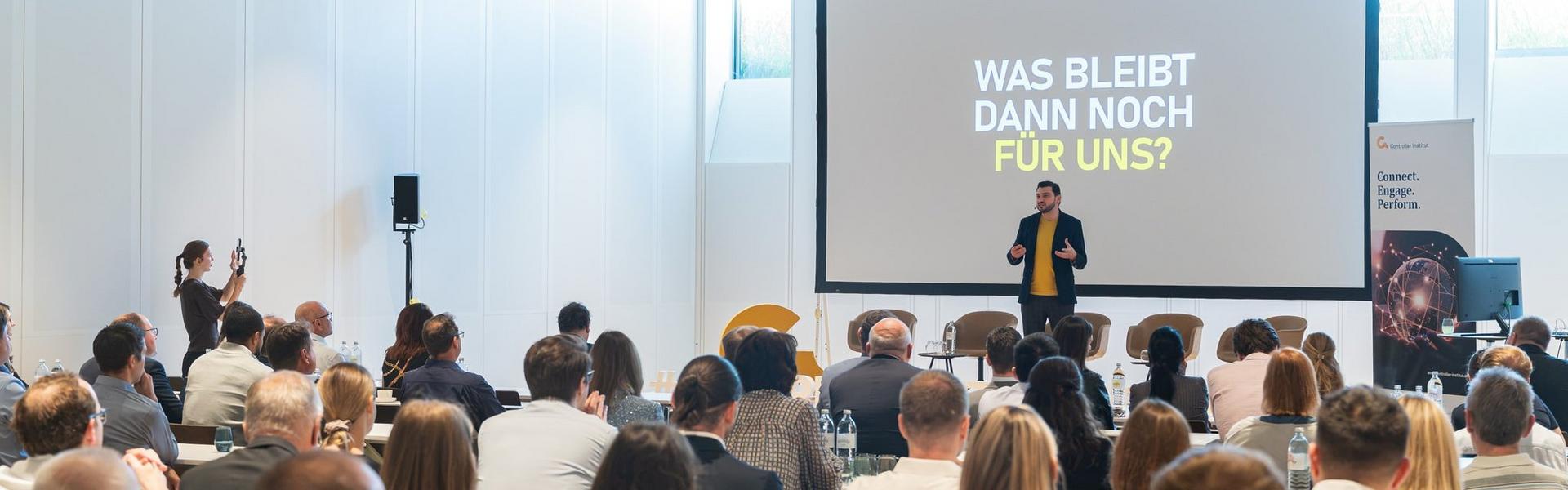 Konferenzraum mit Publikum und Vortragendem vor einer Leinwand mit Text