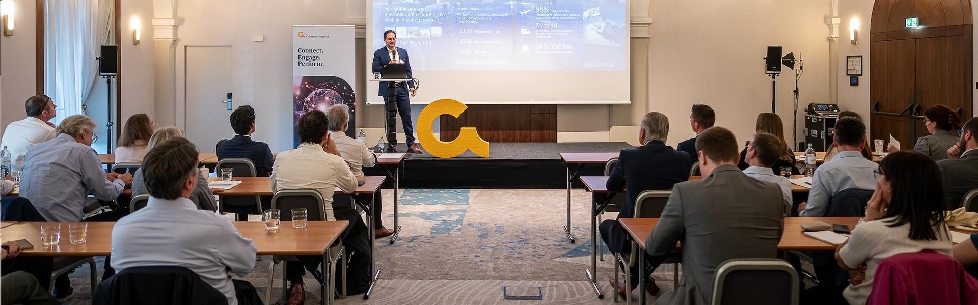 Konferenzraum mit Publikum, das einem Redner auf einer Bühne mit großem Bildschirm und gelbem Buchstaben-C zuhört