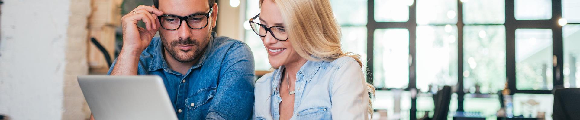 Zwei Personen sitzen an einem Tisch und schauen gemeinsam auf einen Laptop in einem hellen, modernen Büro mit großen Fenstern Zwei Personen sitzen an einem Tisch und schauen gemeinsam auf einen Laptop in einem hellen, modernen Büro mit großen Fenstern