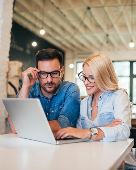 Zwei Personen sitzen an einem Tisch und schauen gemeinsam auf einen Laptop in einem hellen, modernen Büro mit großen Fenstern