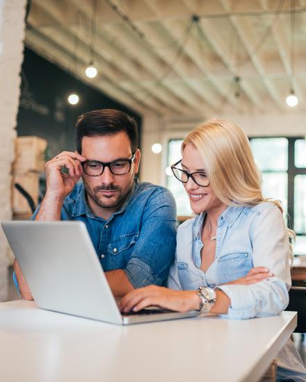 Zwei Personen sitzen an einem Tisch und schauen gemeinsam auf einen Laptop in einem hellen, modernen Büro mit großen Fenstern