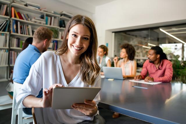 Junge Frau lächelt und hält Tablet, im Hintergrund arbeiten vier Personen an einem Tisch in einem modernen Büro