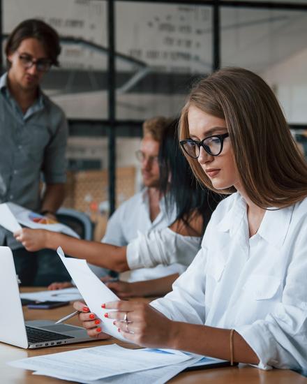 Zwei Personen in einem Büro, eine sitzt am Tisch und betrachtet Papiere, die andere steht dahinter und hält ein Tablet