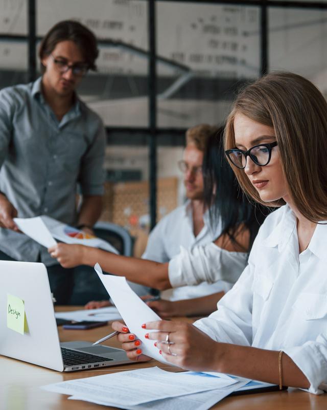 Zwei Personen in einem Büro, eine sitzt am Tisch und betrachtet Papiere, die andere steht dahinter und hält ein Tablet Zwei Personen in einem Büro, eine sitzt am Tisch und betrachtet Papiere, die andere steht dahinter und hält ein Tablet