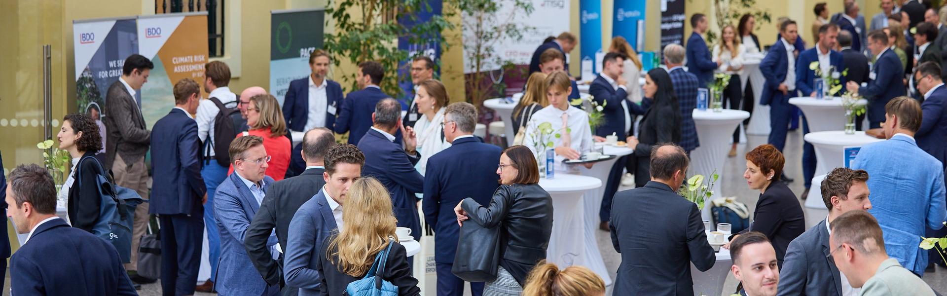 Austrian CFO Day 2025 Mehrere Personen in Businesskleidung stehen an Stehtischen und unterhalten sich auf einer Veranstaltung in einem hellen Innenhof mit gelben Wänden und großen Fenstern.