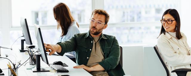 Drei Personen arbeiten in einem hellen Büro an Computern, zwei sitzen nebeneinander und eine Person arbeitet im Hintergrund.