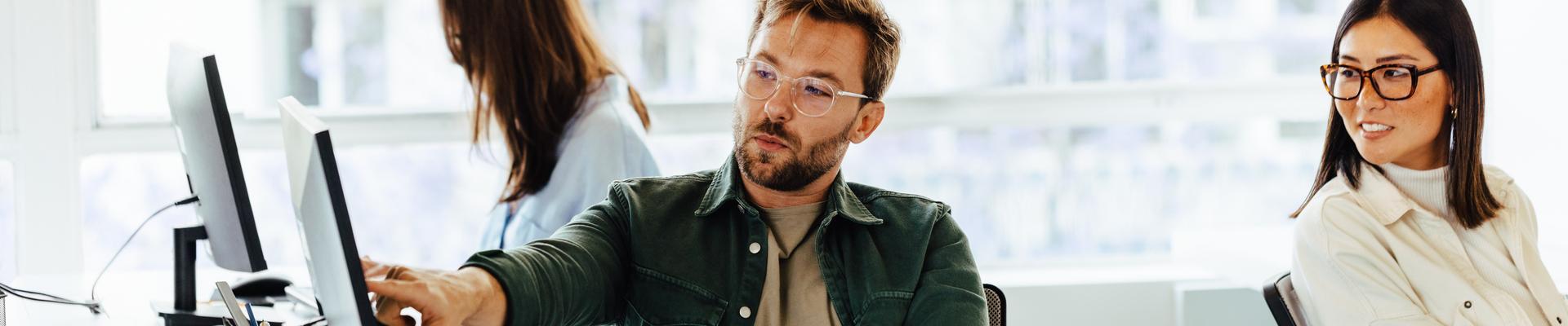 Drei Personen arbeiten in einem hellen Büro an Computern, zwei sitzen nebeneinander und eine Person arbeitet im Hintergrund.