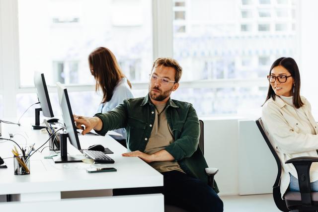 Drei Personen arbeiten in einem hellen Büro an Computern, zwei sitzen nebeneinander und eine Person arbeitet im Hintergrund.
