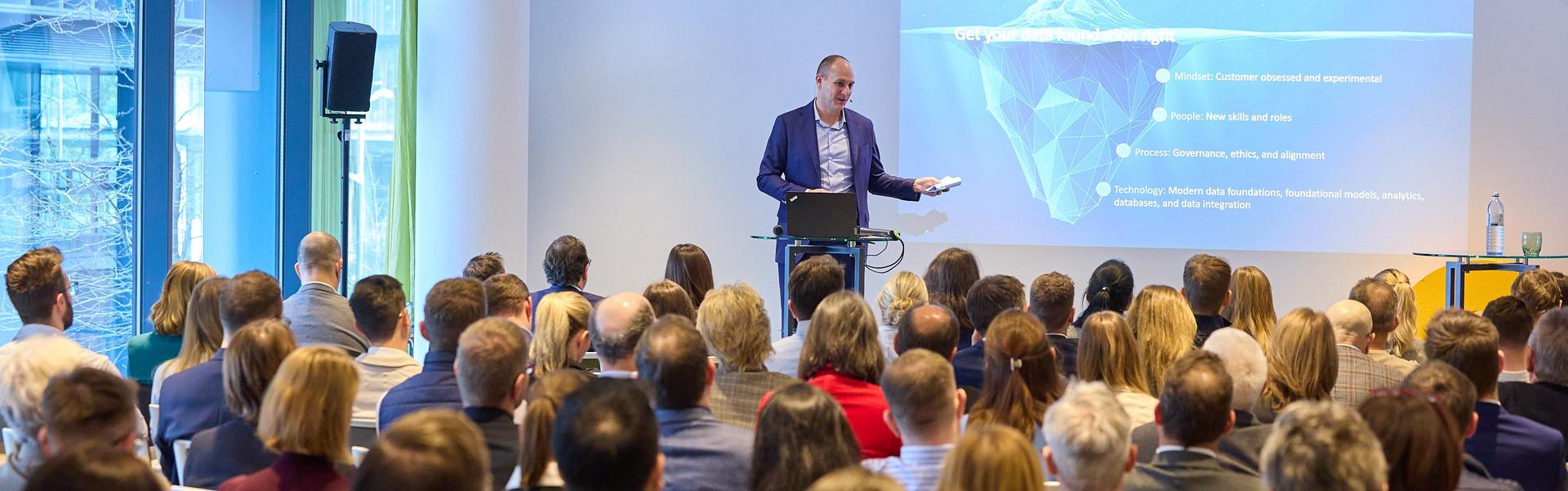 Ein Mann steht vor einem Publikum und hält eine Präsentation mit einer Folie, die ein Eisberg-Diagramm zeigt.