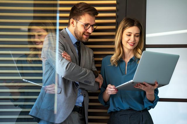Zwei Kollegen, ein Mann im Anzug und eine Frau mit blauem Hemd, schauen gemeinsam auf einen Laptop